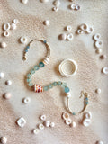 A beaded necklace with light blue-green glass beads, a striped shell charm, and cream-colored beads, arranged in a curved shape on a sandy surface surrounded by small seashells.