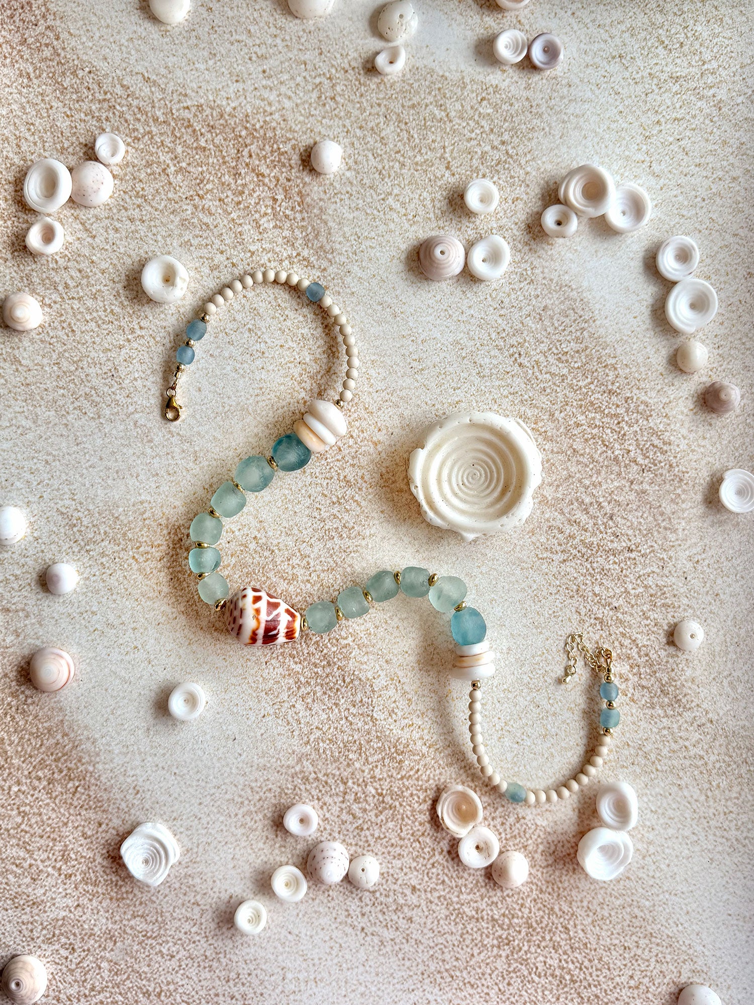 A beaded necklace with light blue-green glass beads, a striped shell charm, and cream-colored beads, arranged in a curved shape on a sandy surface surrounded by small seashells.