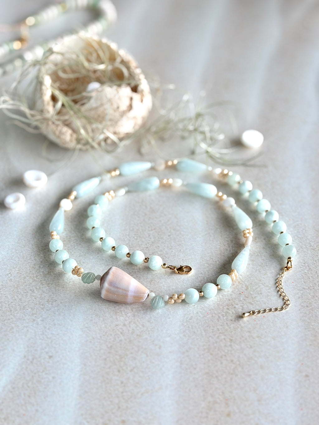 A delicate beaded necklace with pale blue-green beads, small gold spacer beads, and cone shell accent, displayed on a textured light beige surface with boho-style decorative props.