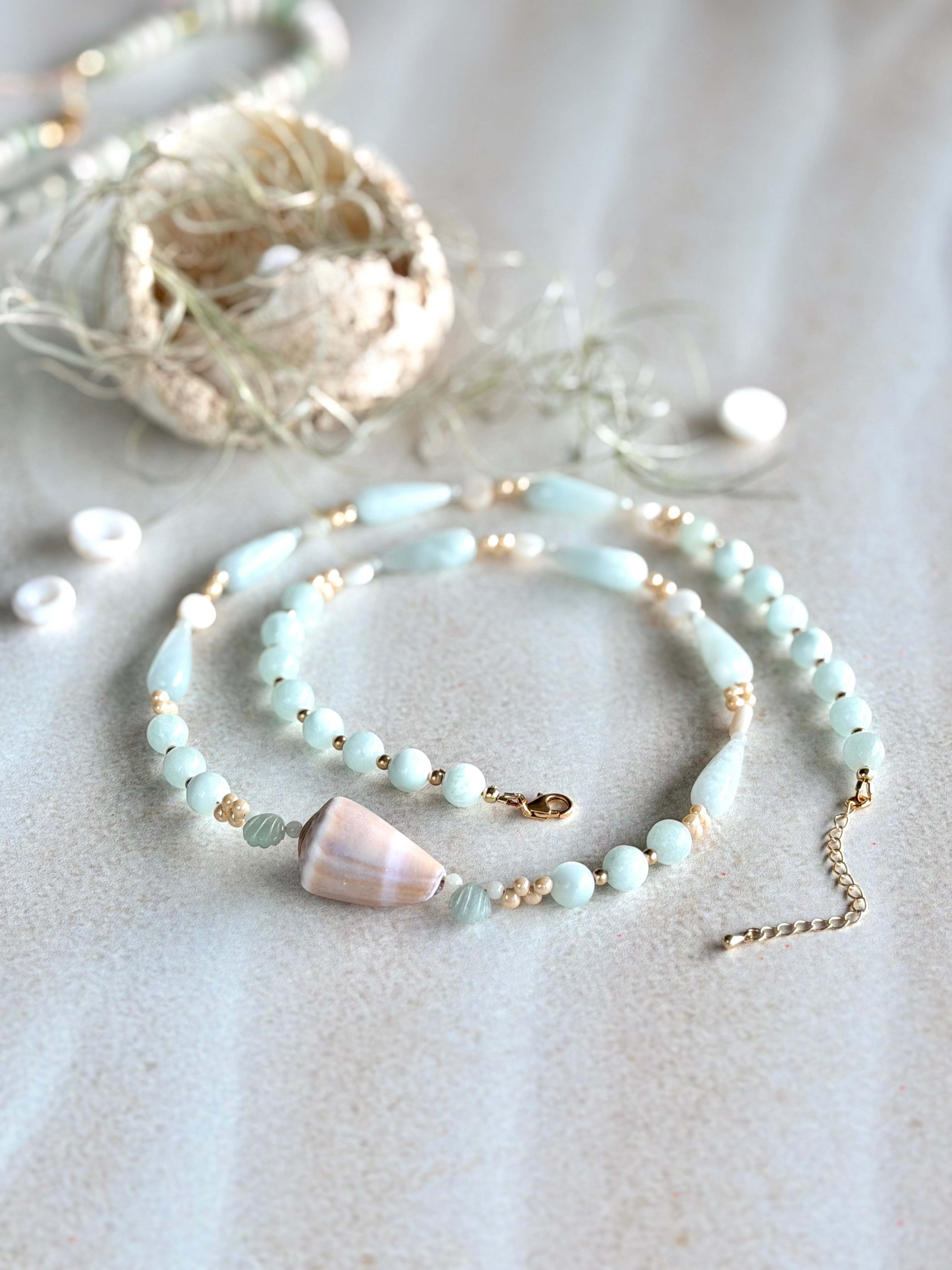A delicate beaded necklace with pale blue-green beads, small gold spacer beads, and cone shell accent, displayed on a textured light beige surface with boho-style decorative props.