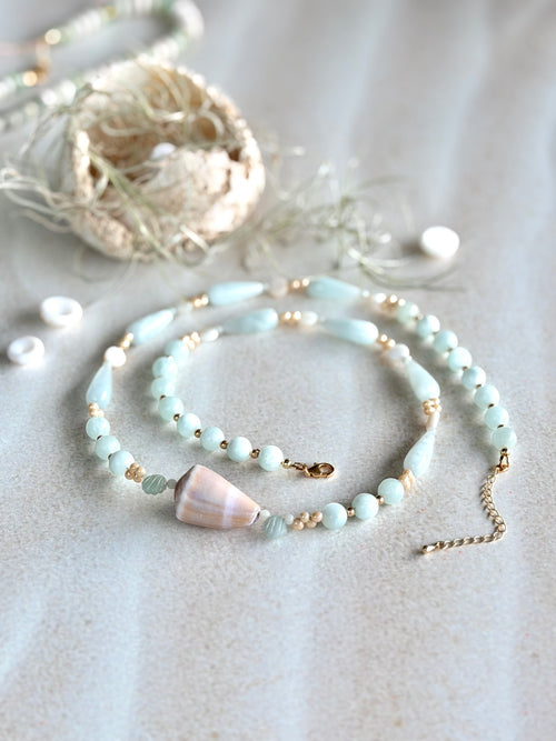 A delicate beaded necklace with pale blue-green beads, small gold spacer beads, and cone shell accent, displayed on a textured light beige surface with boho-style decorative props.