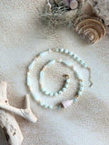 A beaded necklace and matching bracelet in pale aqua and light pink, styled on a sandy surface with seashells, driftwood, and sea plants.