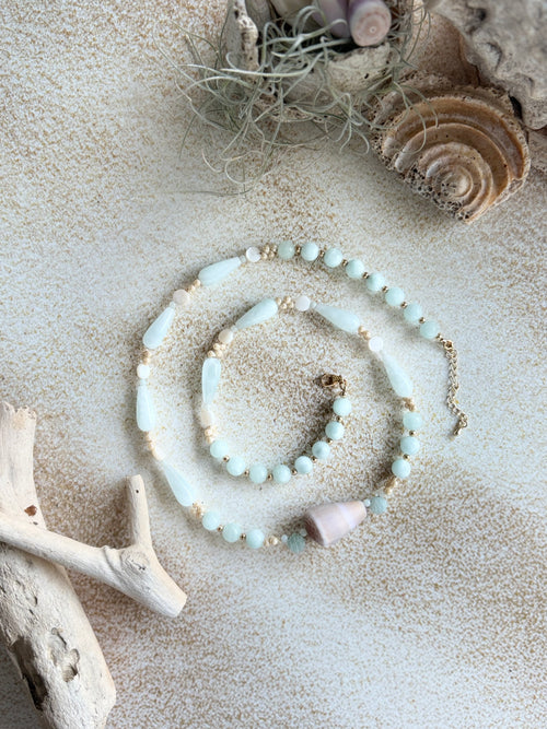 A beaded necklace and matching bracelet in pale aqua and light pink, styled on a sandy surface with seashells, driftwood, and sea plants.