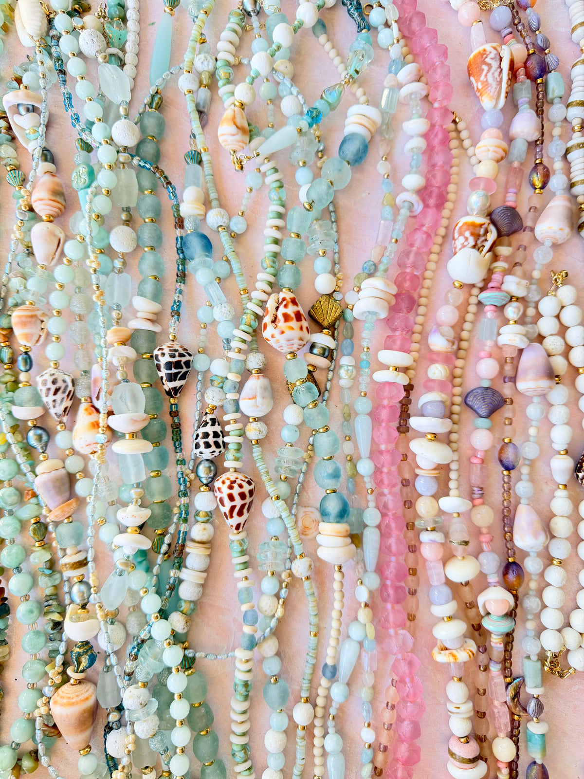 A close-up of multiple strands of beaded necklaces hanging vertically, featuring pastel aqua, mint, pink, cream, and white beads with small seashell and wooden bead accents against a soft pink background.