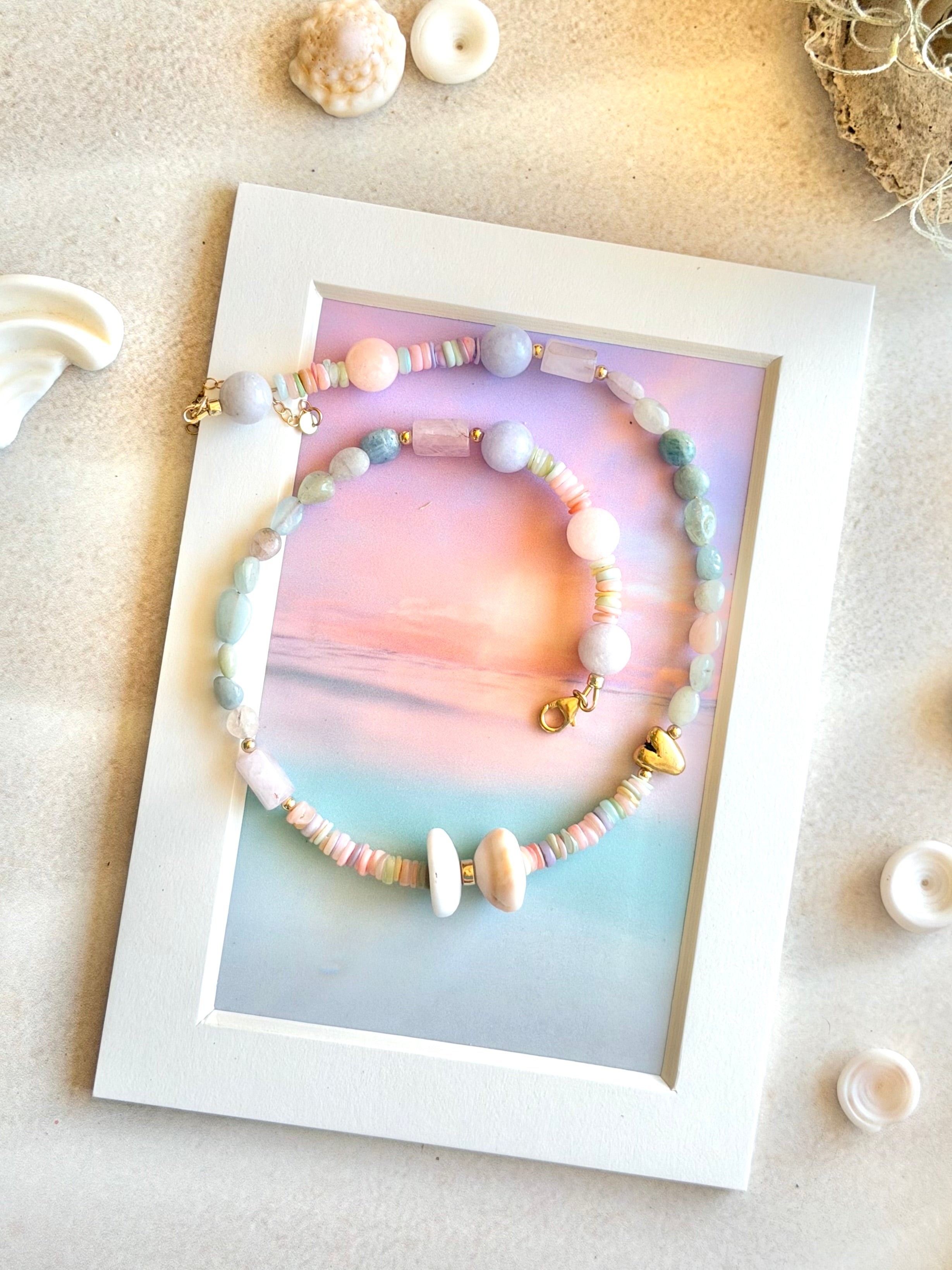 pastel beaded necklace displayed on a white picture frame over a soft sunset print. The bracelets feature pastel pink, blue, green, and lavender beads with small gold charms, including a heart charm and a tiny shell charm.