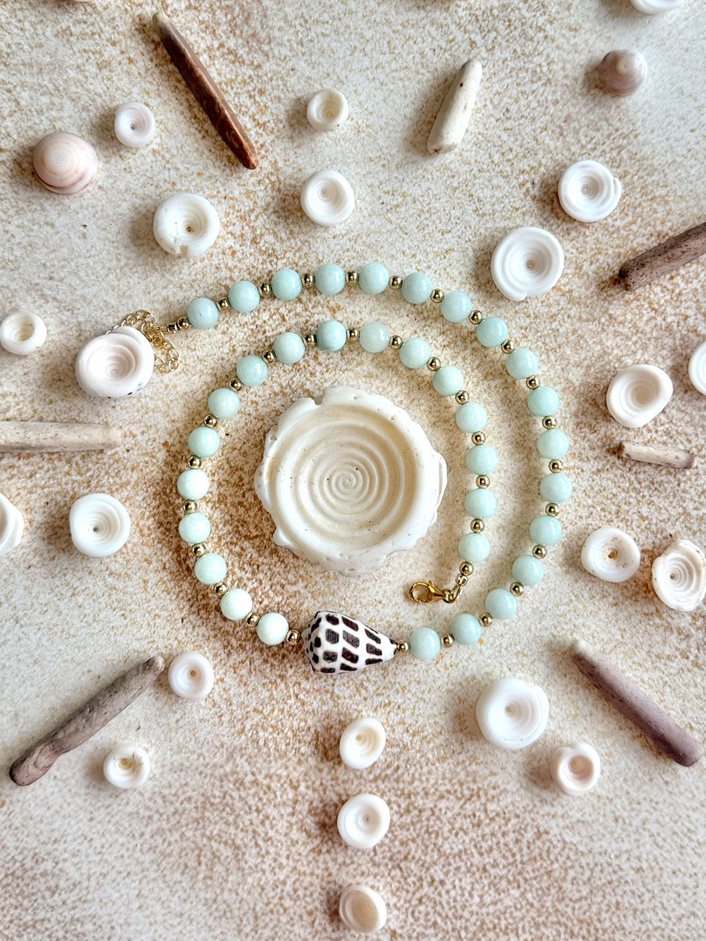 A beaded bracelet arranged in a spiral on a sandy surface, surrounded by small seashells and driftwood. The bracelet features pale mint green beads, small gold spacer beads, and a central white spiral shell charm with a black-and-white patterned accent bead nearby.