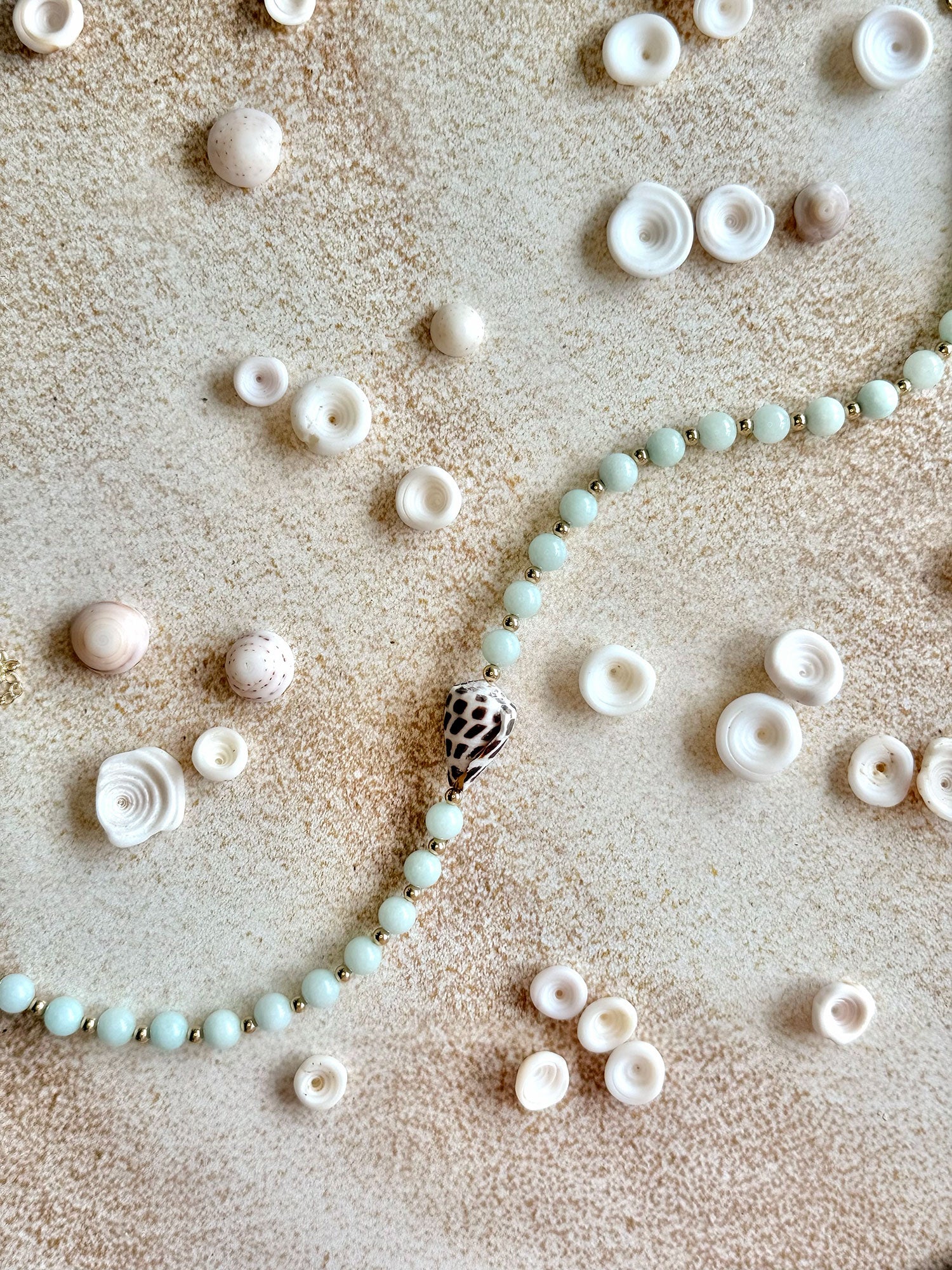A beaded strand with pale mint-green round beads and a central spiral shell pendant, arranged on a textured beige surface with small white and pink seashells scattered around.