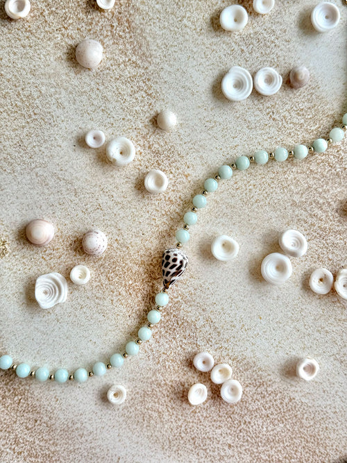 A beaded strand with pale mint-green round beads and a central spiral shell pendant, arranged on a textured beige surface with small white and pink seashells scattered around.