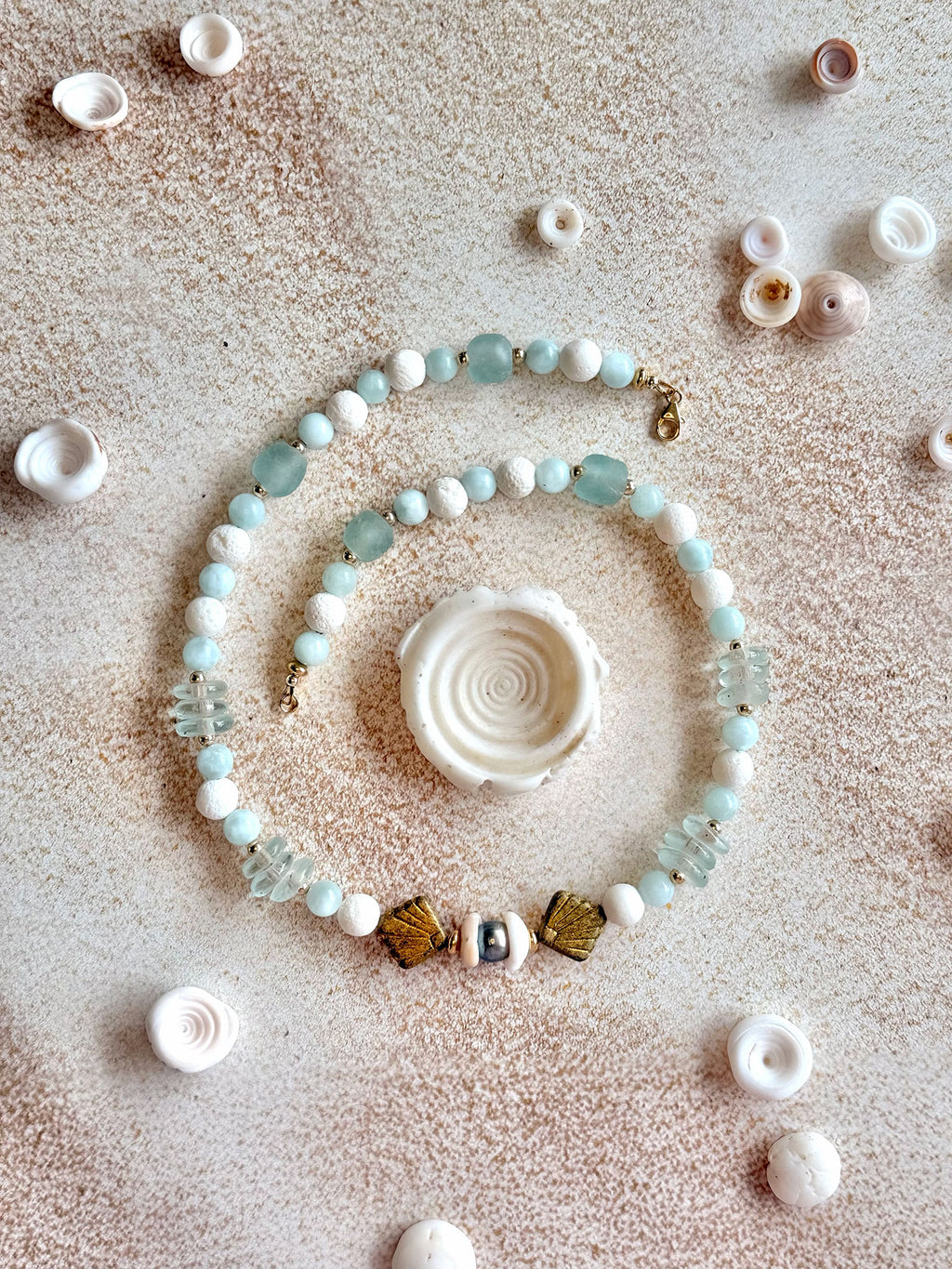 A beaded strand jewelry piece arranged in a spiral on a textured beige surface, surrounded by small seashells. The beads are primarily light blue-green, white, and gold-toned, with a central white spiral shell.