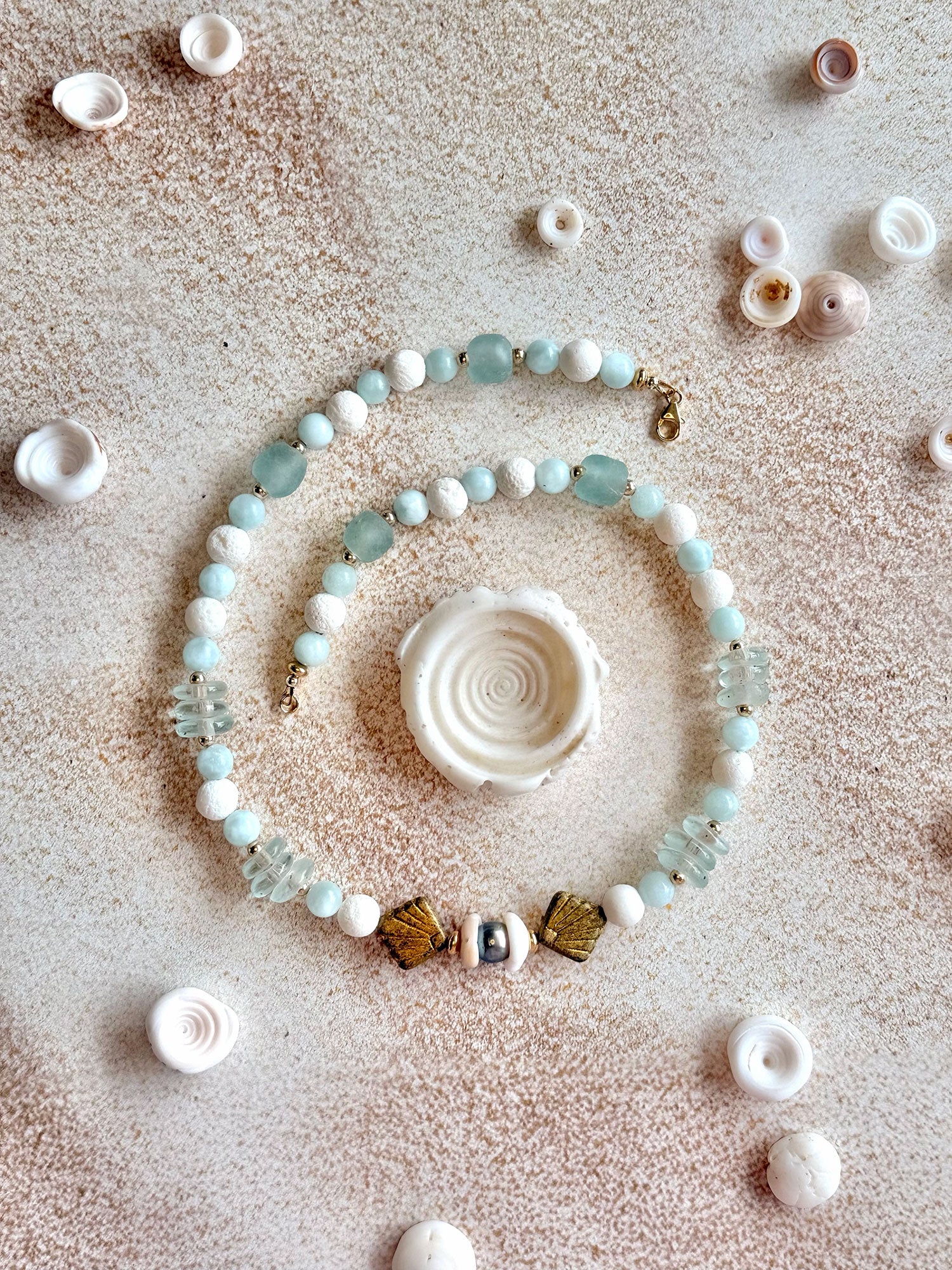 A beaded strand jewelry piece arranged in a spiral on a textured beige surface, surrounded by small seashells. The beads are primarily light blue-green, white, and gold-toned, with a central white spiral shell.