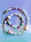 A beaded necklace on a pastel purple and blue background. The jewelry features white beads, light green cylindrical beads, lavender-purple shell-like beads, and large cowrie shells in soft pink, cream, and lavender.
