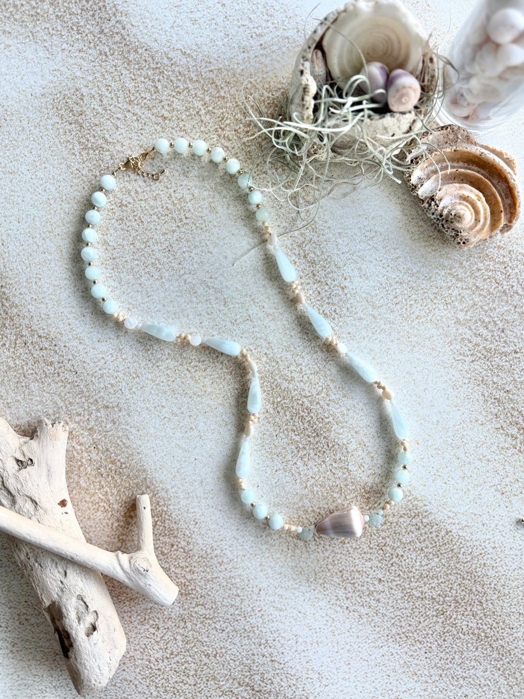A beaded necklace with pale blue-green beads and small gold-toned accents, arranged on a sandy surface with seashells, driftwood, and sea plants as styled props.