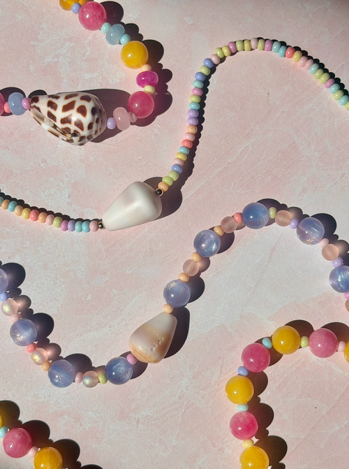 beaded necklaces with pastel rainbow beads and large white teardrop-shaped beads, arranged on a pink textured background with a seashell accent.