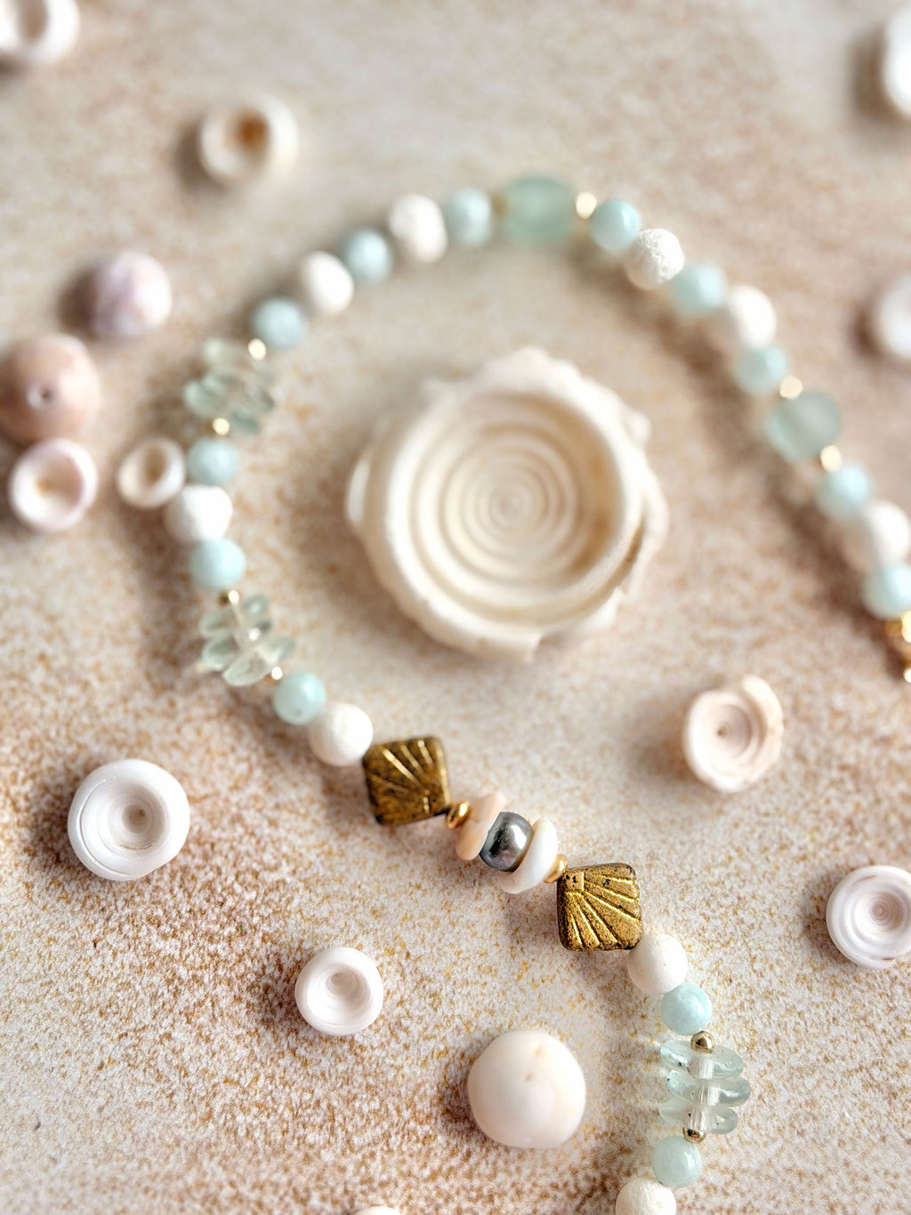 A beaded strand necklace or bracelet laid on a sandy surface with small seashells and pale stones around it. The strand features white beads, pale aqua beads, clear faceted beads, and two metallic gold shell-shaped beads.