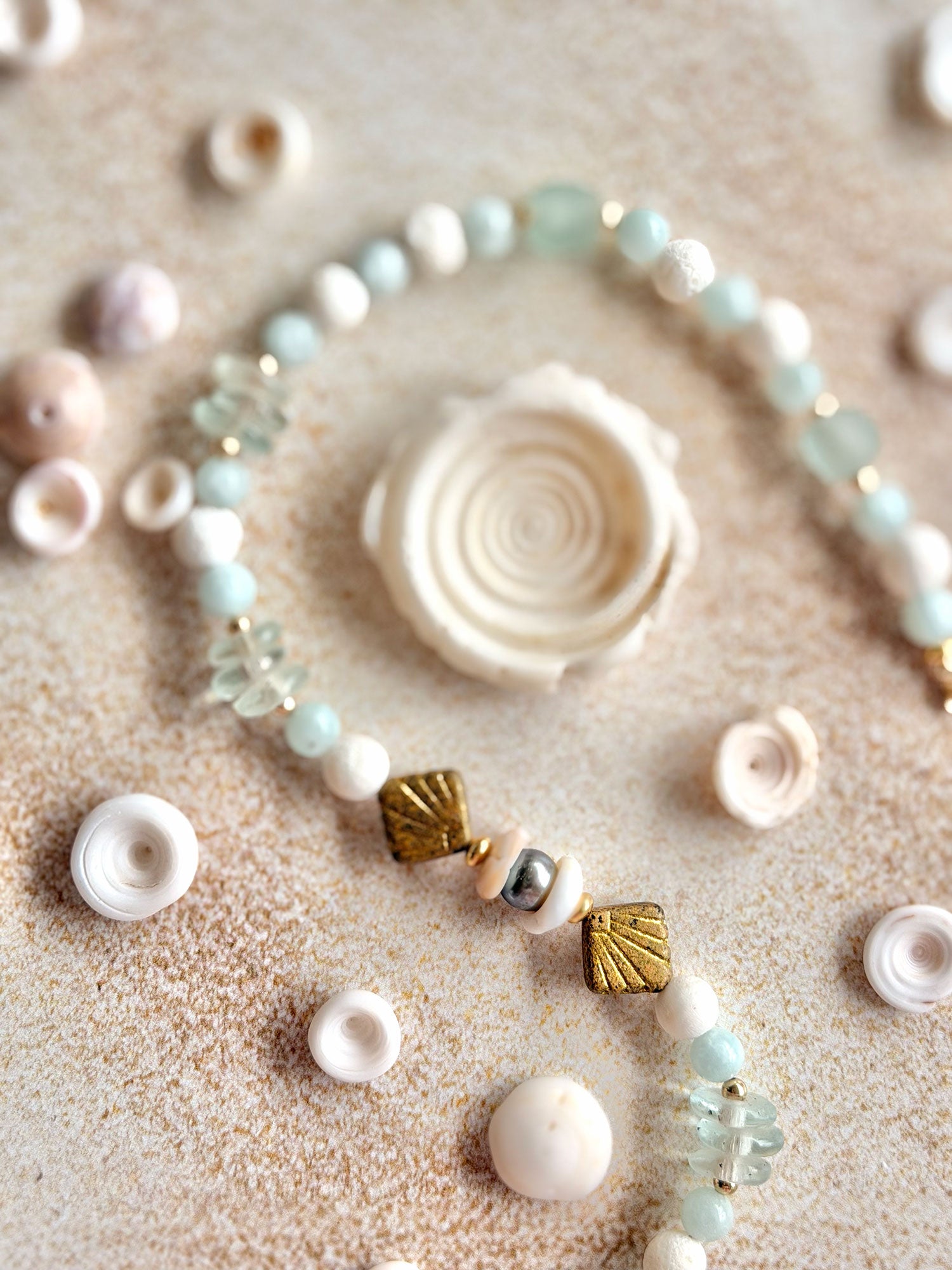 A beaded strand necklace or bracelet laid on a sandy surface with small seashells and pale stones around it. The strand features white beads, pale aqua beads, clear faceted beads, and two metallic gold shell-shaped beads.