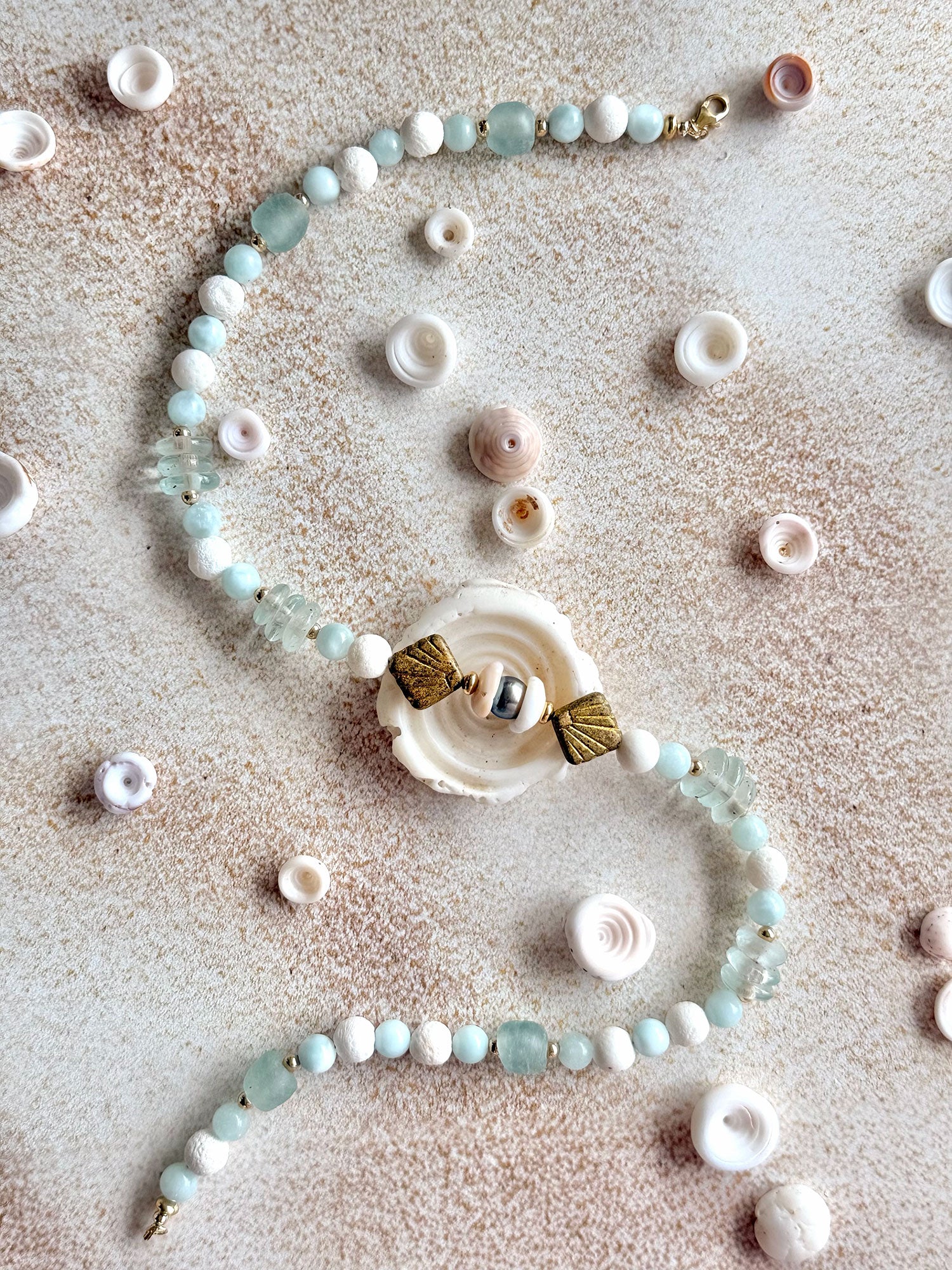 A handmade beaded necklace with light blue and white beads, a central white shell charm, and gold-toned accents, displayed on a textured beige surface with scattered seashells.